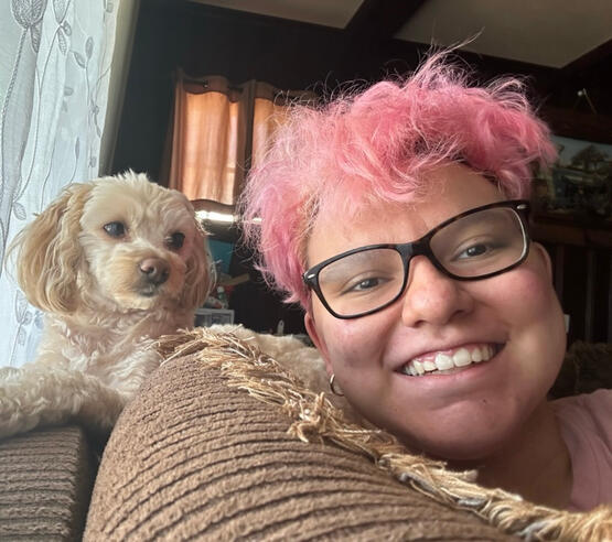 A picture of Timothy Rafael with their dog, Sunny. Tim has light skin, short, curly pink hair, and wears black glasses. He smiles at the camera, while Sunny, a blonde, fluffy maltipoo, stares at him